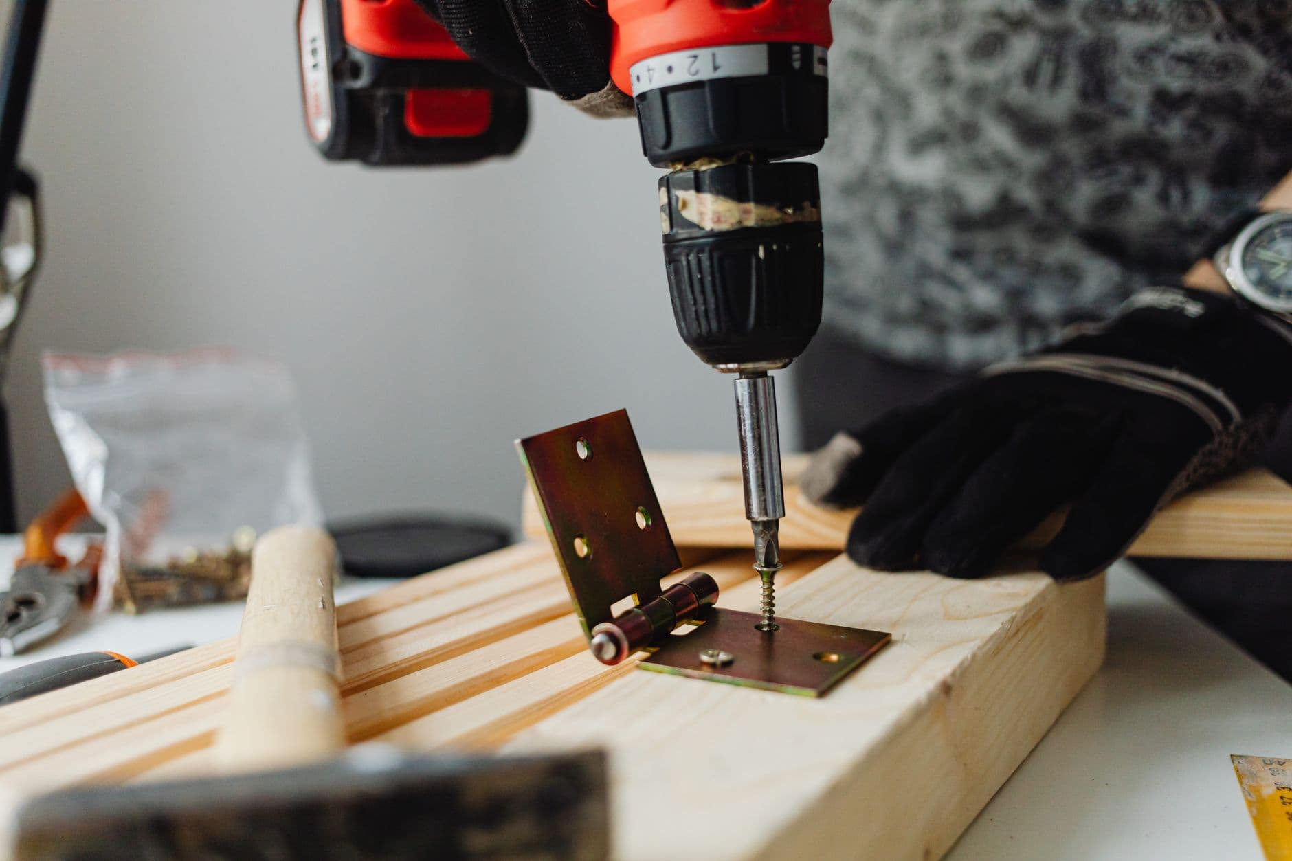 A cordless drill driving a screw through a metal hinge into a length of timber, with a hammer and loose screws alongside.